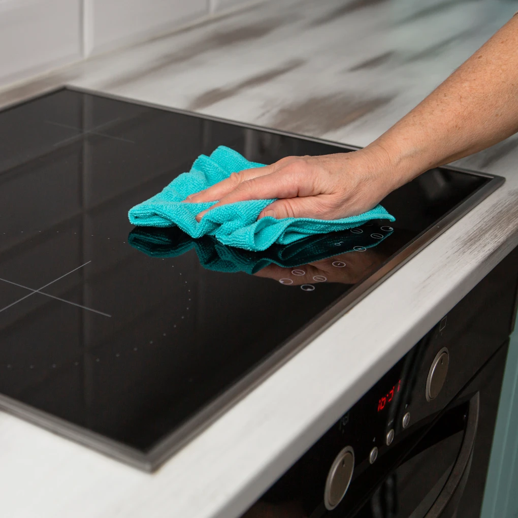 A woman's hand, using a blue microfiber cloth, rubs the glass ceramic stove in the kitchen.